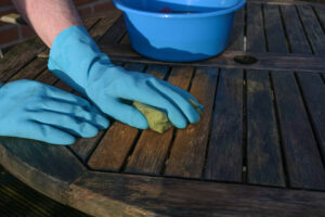 Hände in blauen Gummihandschuhen reinigen zu Beginn des Frühlings einen verwitterten Gartentisch aus Holz für die Outdoor-Saison